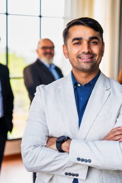 Closeup portrait of young Indian Asian business professional or CEO standing confidently with folded hands and a smile, colleagues blurred in the background at a corporate office